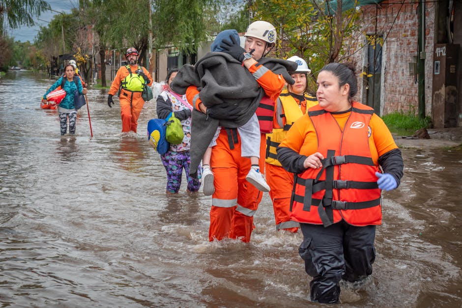 A group of emergency rescue personnel wearing bright orange waterproof suits and life vests wading through a flooded street during a rescue operation. One rescuer is carrying a distressed child wrappe