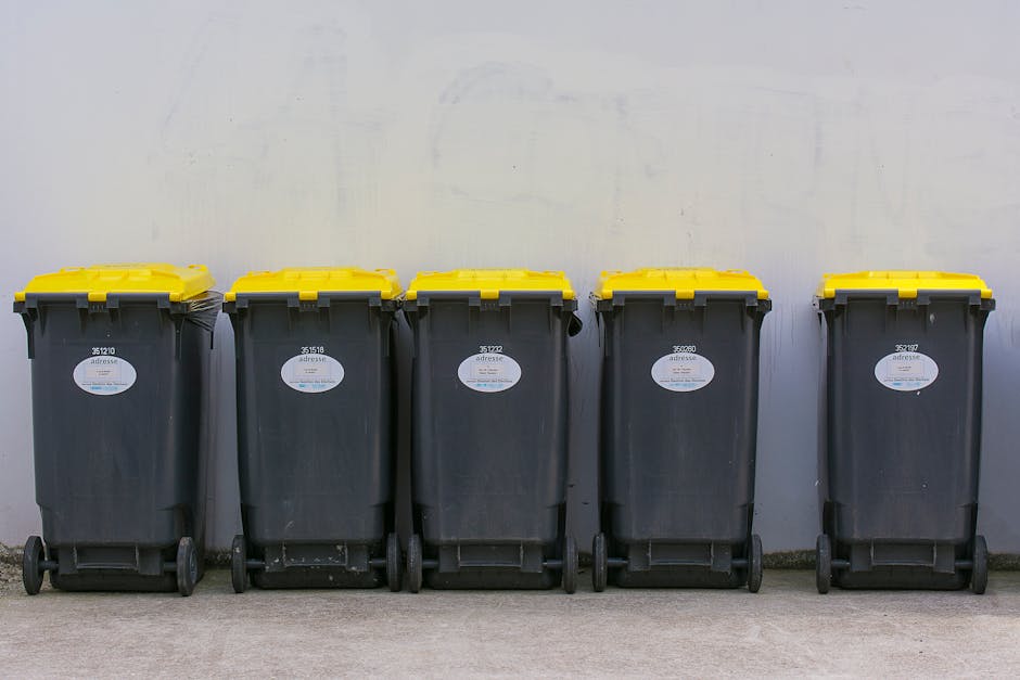 An overflowing outdoor waste disposal area with multiple garbage bins filled with mixed recyclables, black rubbish bags, and cardboard boxes scattered on the ground. Behind the bins, a commercial buil